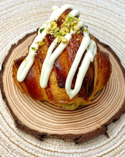 Cruasán casero hecho por la Pilareta, relleno de crema de pistacho y por encima chocolate blanco acompañado de trocitos de pistacho en la parte superior.