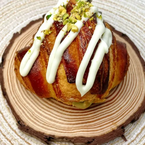 Cruasán casero hecho por la Pilareta, relleno de crema de pistacho y por encima chocolate blanco acompañado de trocitos de pistacho en la parte superior.